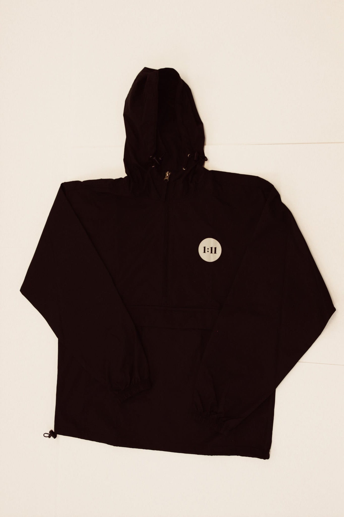 A black hooded jacket or windbreaker laid flat on a light off-white background. The hood is extended to the right, and a small circular white tag with black text is visible on the chest.