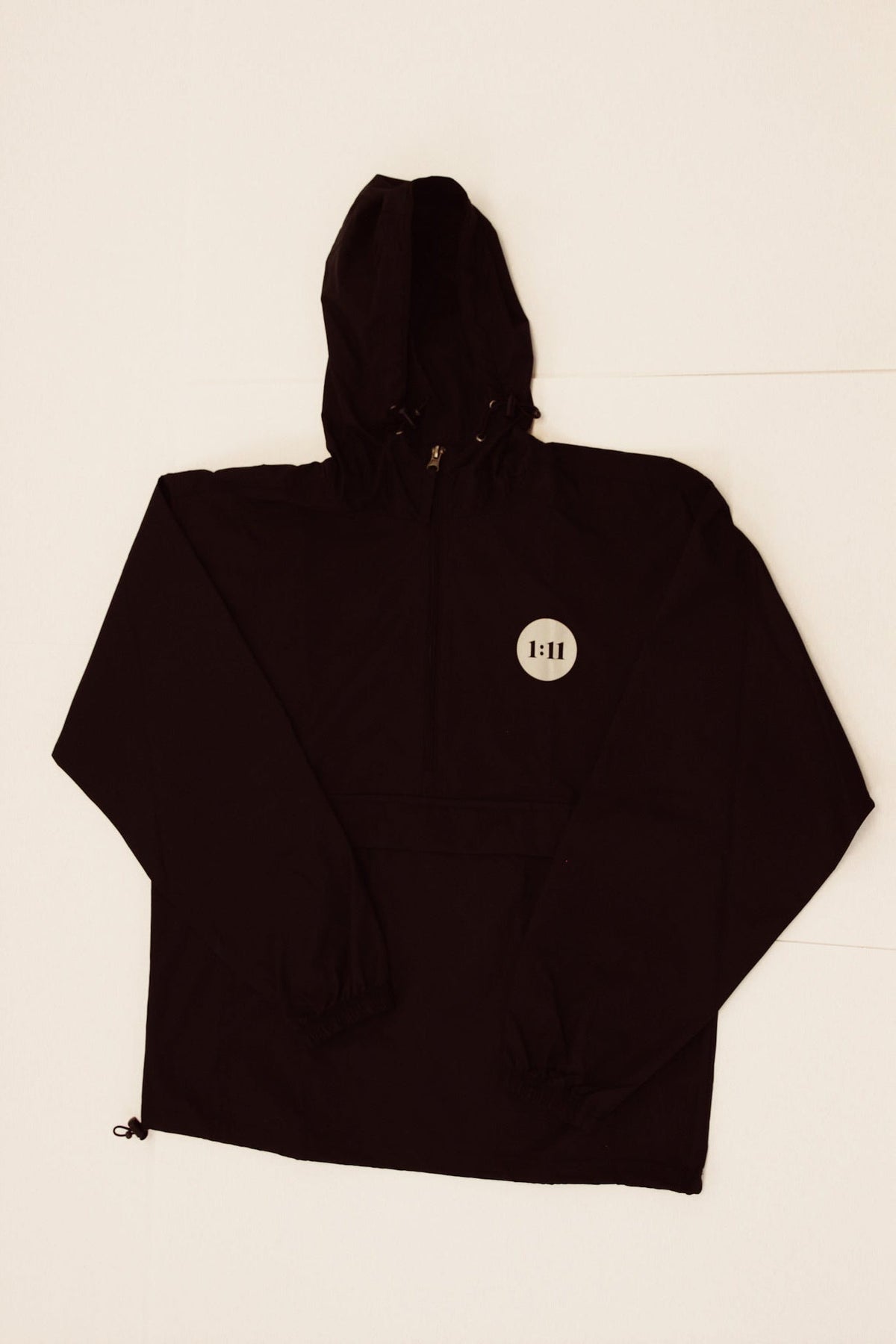 A black hooded jacket or windbreaker laid flat on a light off-white background. The hood is extended to the right, and a small circular white tag with black text is visible on the chest.